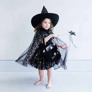 Little girl in a Mimi & Lula Aurora Witch Cape, wearing a black witch hat and holding a magic wand with a cute cat-shaped bag.