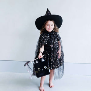 Child in sparkly black witch costume with hat and cape from Mimi & Lula, holding a cute cat-shaped bag. Perfect for Halloween magic!