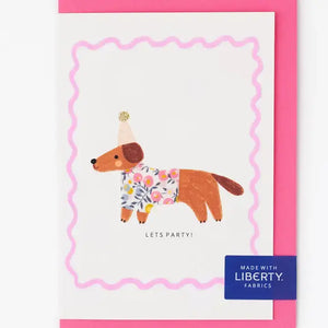 Sausage dog birthday card wearing Liberty fabric Wiltshire bud coat with a golden glitter party hat and "Let's Party" message.