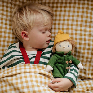 A sleeping child in striped pajamas cuddles Charlie Chestnut, a plush doll in a green coat and yellow beanie, amidst cozy bedding.