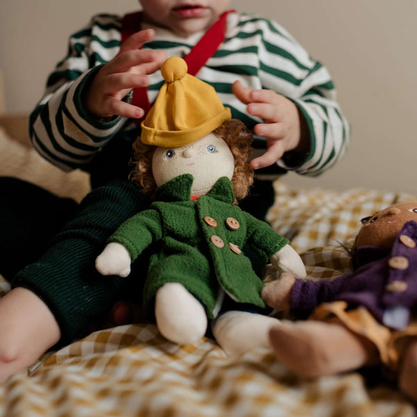 Child playing with Olli Ella Dress Me Dinky Charlie Chestnut doll in green coat and yellow beanie on a cozy bed.