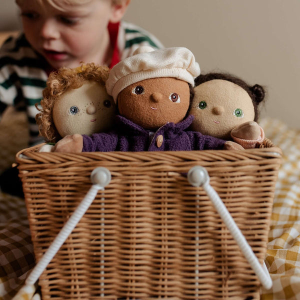 Three adorable dolls peek out of a wicker basket while a child plays in the background, ready for imaginative adventures.