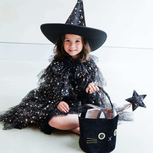 Little girl wearing Mimi & Lula Aurora Witch Hat with starry black costume, holding a cat-shaped bag and star wand, ready for Halloween magic.