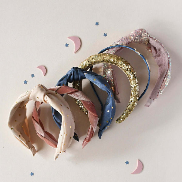 Assorted sequin and knotted headbands in pink, gold, and blue on a soft background with moon and star decorations.